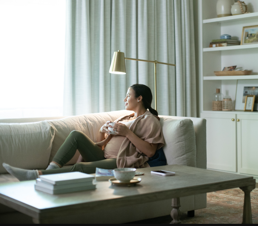 A pregnant woman sitting on a couch looking out a window drinking coffee