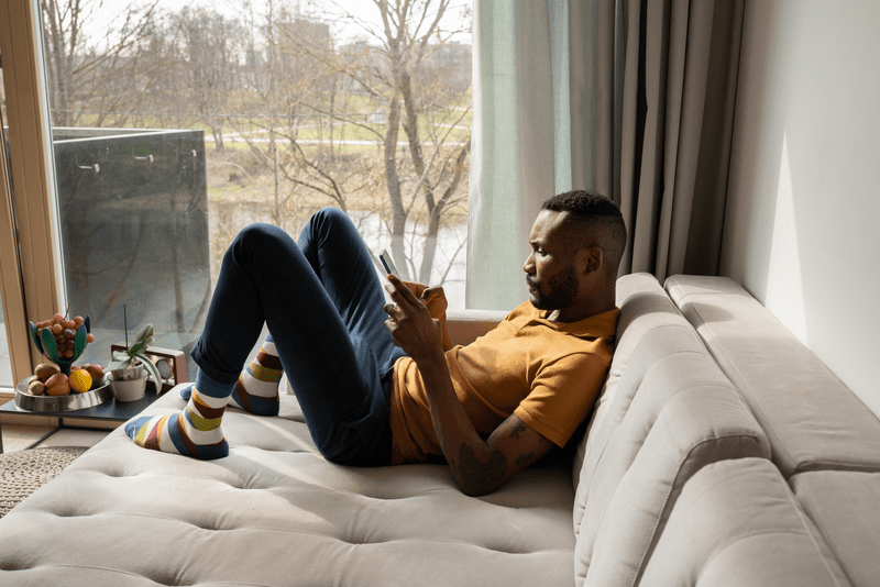Stock Photo Of Man Scrolling Smartphone While Laying On Sofa.